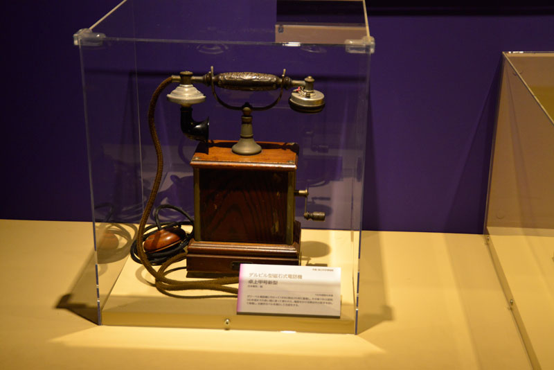 デルビル型磁石式電話機 卓上甲号新型 日本電気製　1929（昭和4）年頃 ガワーベル電話機に代わって1896（明治29）年に登場し、その後1963（昭和38）年頃までの長い間に渡って使われた。電話をかけるときは外の取手を回して発電し、交換手のベルを鳴らして合図する。交換手は口頭で電話番号を聞くと、手動で先方に繋ぐ。 とあります。ガワーベル電話機とはでかいイヤホンのようなものをてで耳に押し当て、電話機に備え付けられた送話口に向かって話す、国産第一号機のようなタイプの電話機で、このように送話器と受話器がハンドセットになっている現在の電話機のようなものではありませんでした。よく考えたらこのデルビル型、ダイヤルはないですけど今の電話と基本的に変わっていないですものね。